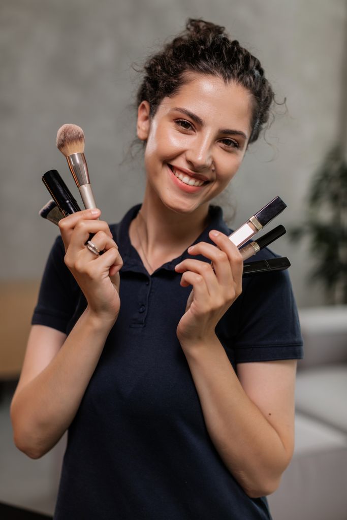 Cheerful woman in polo shirt holding makeup brushes, showcasing beauty products with a smile.